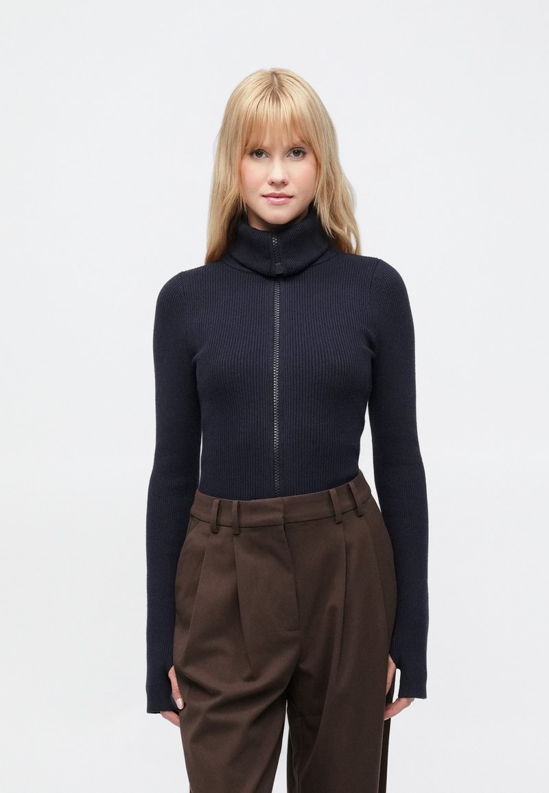 Maglione dolcevita a coste blu navy con zip frontale completa, maniche lunghe e vestibilità aderente, abbinato a pantaloni marrone chiaro a gamba larga.