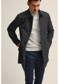 Musta kevyt takki, jossa on kaulus, nappikiinnitys edessä ja slim fit -muotoilu; päällä vaaleanharmaa poolo ja tummansiniset housut.