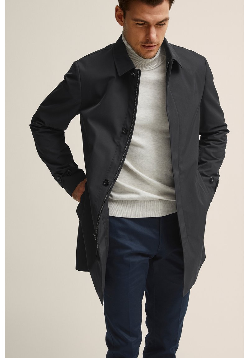 Musta kevyt takki, jossa on kaulus, nappikiinnitys edessä ja slim fit -muotoilu; päällä vaaleanharmaa poolo ja tummansiniset housut.