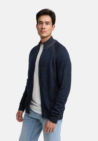 Cardigan zip-up in maglione navy con motivo testurizzato e colletto a coste; indossato sopra una camicia grigia chiaro, abbinato a jeans blu chiaro.