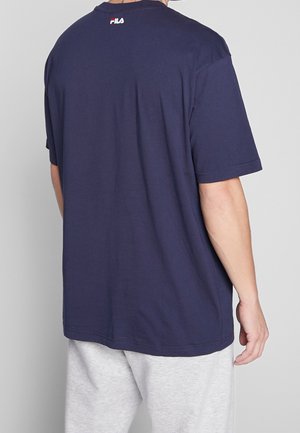 Homme portant un t-shirt FILA bleu marine et un pantalon de survêtement gris clair, vu de dos sur un fond clair et uni.