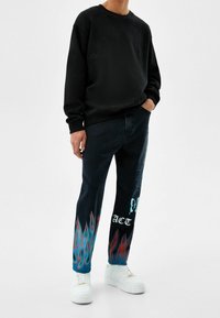 Černá mikina s dlouhými rukávy a klasickým výstřihem, spárovaná s tmavě modrými džíny s plamenovými grafikami v modré a červené barvě, a bílé tenisky.