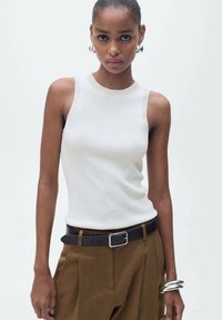 Weißes geripptes Tanktop mit rundem Ausschnitt, kombiniert mit braunen hoch taillierten Hosen und einem schwarzen Gürtel. Silberne Armreifen betonen das Handgelenk.