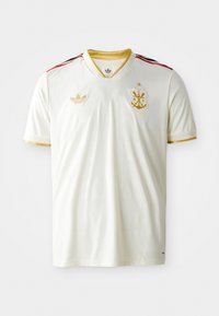 Maglia da calcio bianca con scollatura a V, dettagli dorati, strisce nere e rosse sulle spalle e un emblema dorato sul petto. Texture morbida e liscia.