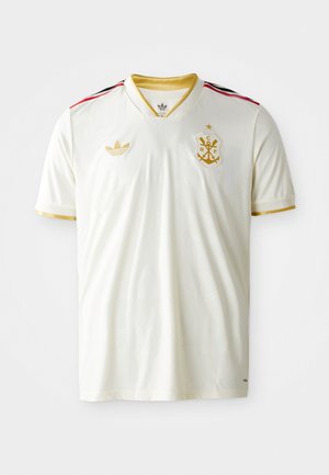 Hvid fodboldtrøje med v-hals, guld accenter, sorte og røde striber på skuldrene, og et guld emblem på brystet. Blød, glat tekstur.