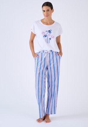 Mujer de pie descalza, con una camiseta blanca de manga corta con estampado de jarrón de flores y pantalones sueltos de rayas azules, rosas y blancas, con las manos en los bolsillos.