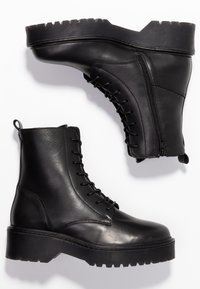 Bottes cheville en cuir noir avec une semelle en caoutchouc texturée, un laçage à l'avant et une fermeture éclair latérale. Elles présentent un bout rond et un talon épais pour plus de stabilité.