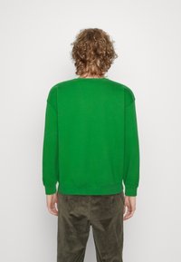 Sudadera verde con cuello redondo, hombros caídos y puños acanalados, combinada con pantalones de pana oliva texturizada.