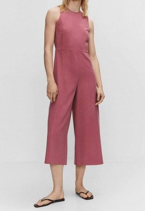 Person iført ærmeløs, bredbenet jumpsuit i støvet rosafarve og sorte flip-flops, stående mod en ensfarvet lys baggrund.