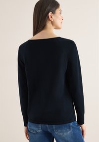 Femme aux longs cheveux bruns portant un haut noir à manches longues et un jean bleu, debout devant un fond clair uni, dos tourné.