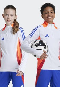 Zwei lächelnde Kinder in weißer und blauer Sportkleidung mit orangefarbenen Akzenten, eines hält einen schwarz-weißen Fußball.