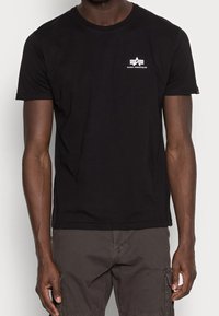 Mężczyzna w czarnej koszulce z krótkim rękawem z małym białym logo Alpha Industries na górnej lewej części klatki piersiowej oraz w ciemnych szortach cargo.
