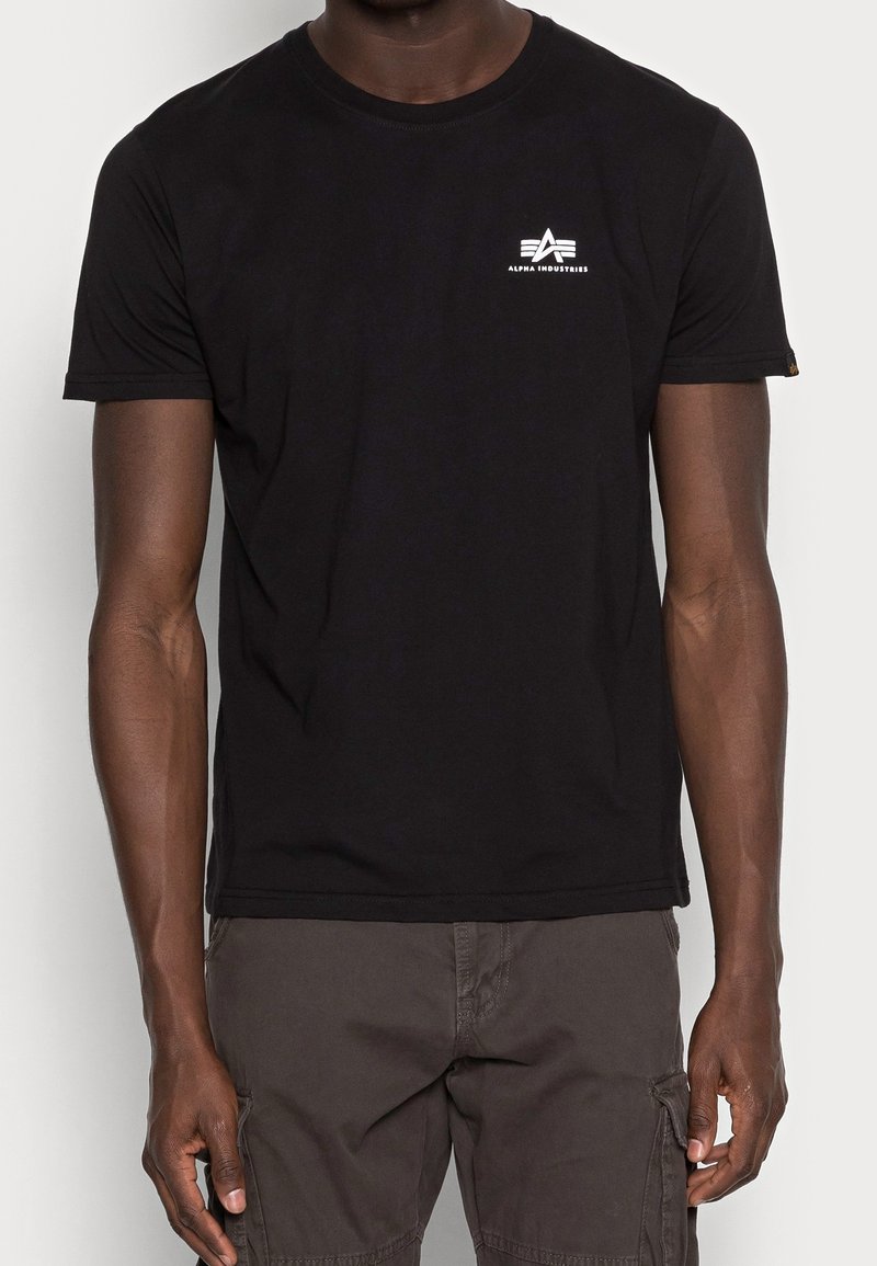 Mężczyzna w czarnej koszulce z krótkim rękawem z małym białym logo Alpha Industries na górnej lewej części klatki piersiowej oraz w ciemnych szortach cargo.