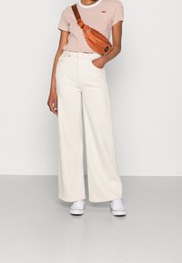 Krämfärgade vida byxor, randig kortärmad topp och en brun crossbody-väska. Vita sneakers. Minimalistisk design, avslappnad stil, med texturerad tyg.