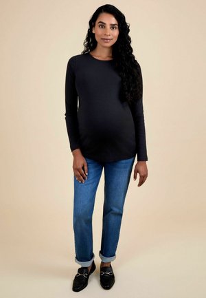 Langärmliges schwarzes Oberteil kombiniert mit hochgekrempelten blauen Jeans und schwarzen Slippern. Glatte Textur; enganliegende Silhouette, die einen runden Bauch betont.