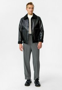 Zwarte leren jas met een schapenvachtkraag, knopenluiting aan de voorkant en twee grote zakken, gecombineerd met grijs gestreepte broek en zwarte schoenen.