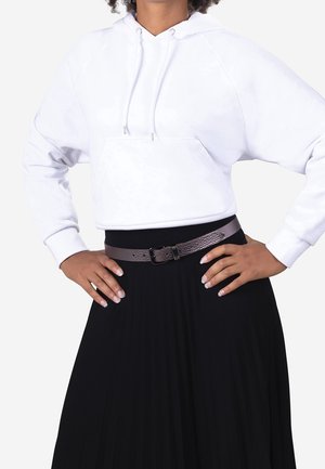 Personne portant un sweat à capuche blanc court, une jupe plissée noire et une ceinture marron texturée, les mains sur les hanches.