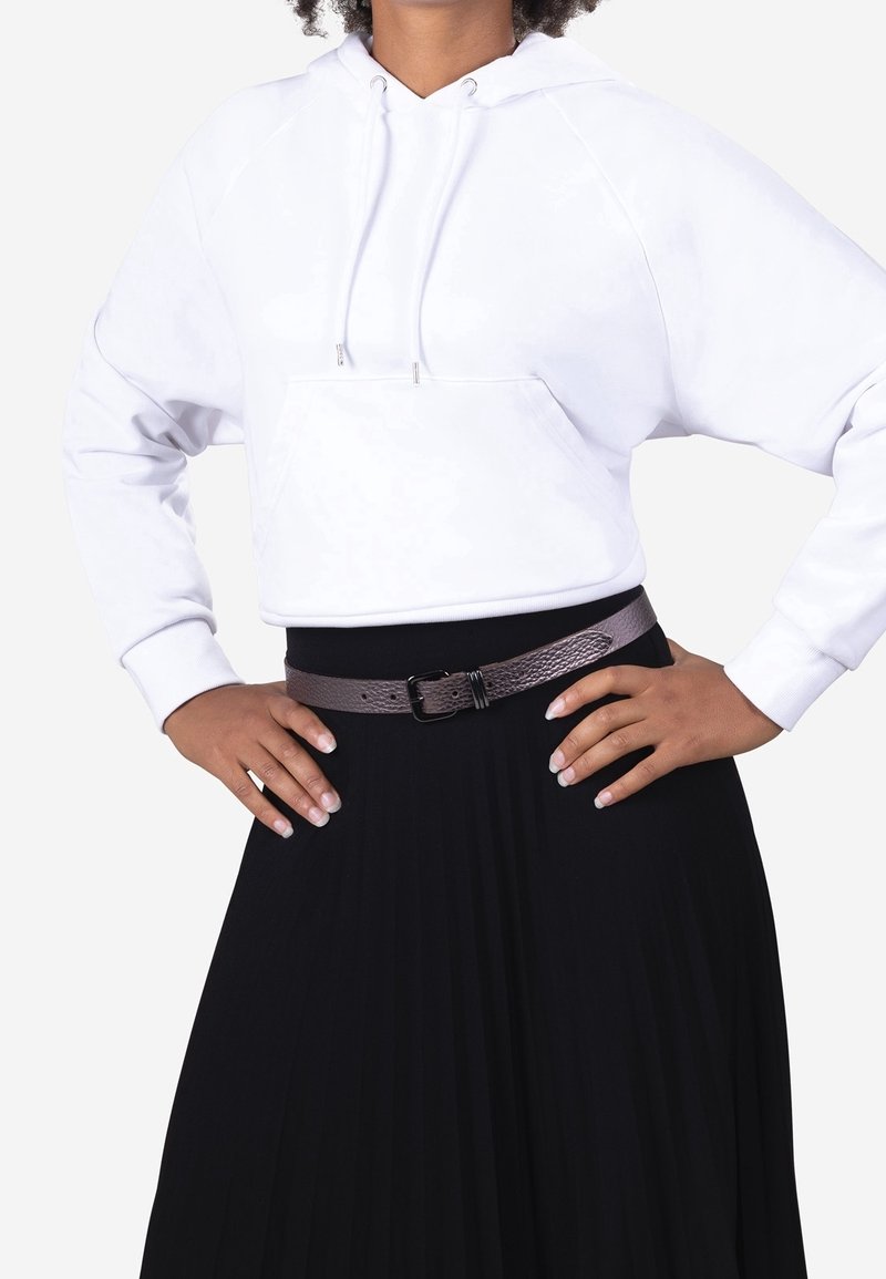 Personne portant un sweat à capuche blanc court, une jupe plissée noire et une ceinture marron texturée, les mains sur les hanches.