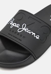 Chancla negra de material de goma texturizado. Presenta un logo embosado prominente y un diseño numérico en la correa. Diseño de suela plana.