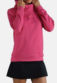 Persona con una sudadera rosa brillante con un pequeño logo, ajustándose el cuello, combinada con una falda negra con pliegues.
