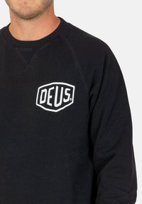 Sudadera negra de algodón, con cuello redondo y puños acanalados. Tiene un logo blanco "DEUS" en el pecho.