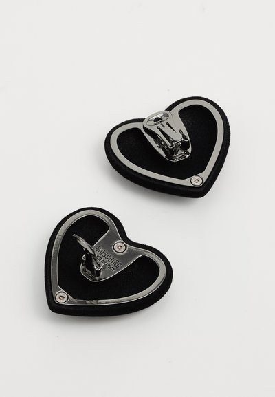Divas melnās sirds formas uzlīmējamas auskaru pāris ar sudraba malām un metāla sprādzēm, viens no tiem ar "Moschino" zīmolu, uz baltas fona.