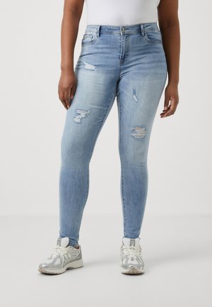 Jean skinny taille haute bleu clair avec des zones déchirées, présentant des déchirures sur les cuisses et une texture lisse, associé à des baskets blanches.