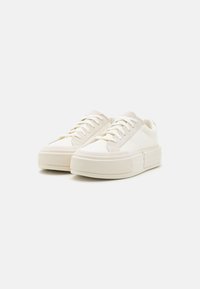 Sneaker bianchi in tela con suola in gomma spessa, lacci piatti e un accento testurizzato sui lati, caratterizzati da un design minimalista.