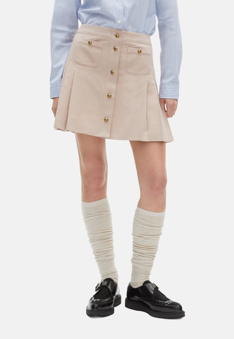 Beige mini-rok met voorzakken en gouden knoopdetails. Geselecteerd met crèmekleurige kniekousen en zwarte schoenen met een chunky zool.