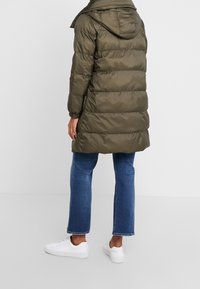 Olivgrön vadderad jacka med huva, horisontell quiltning, midjelång design och elastiska muddar, kombinerad med blå jeans och vita sneakers.