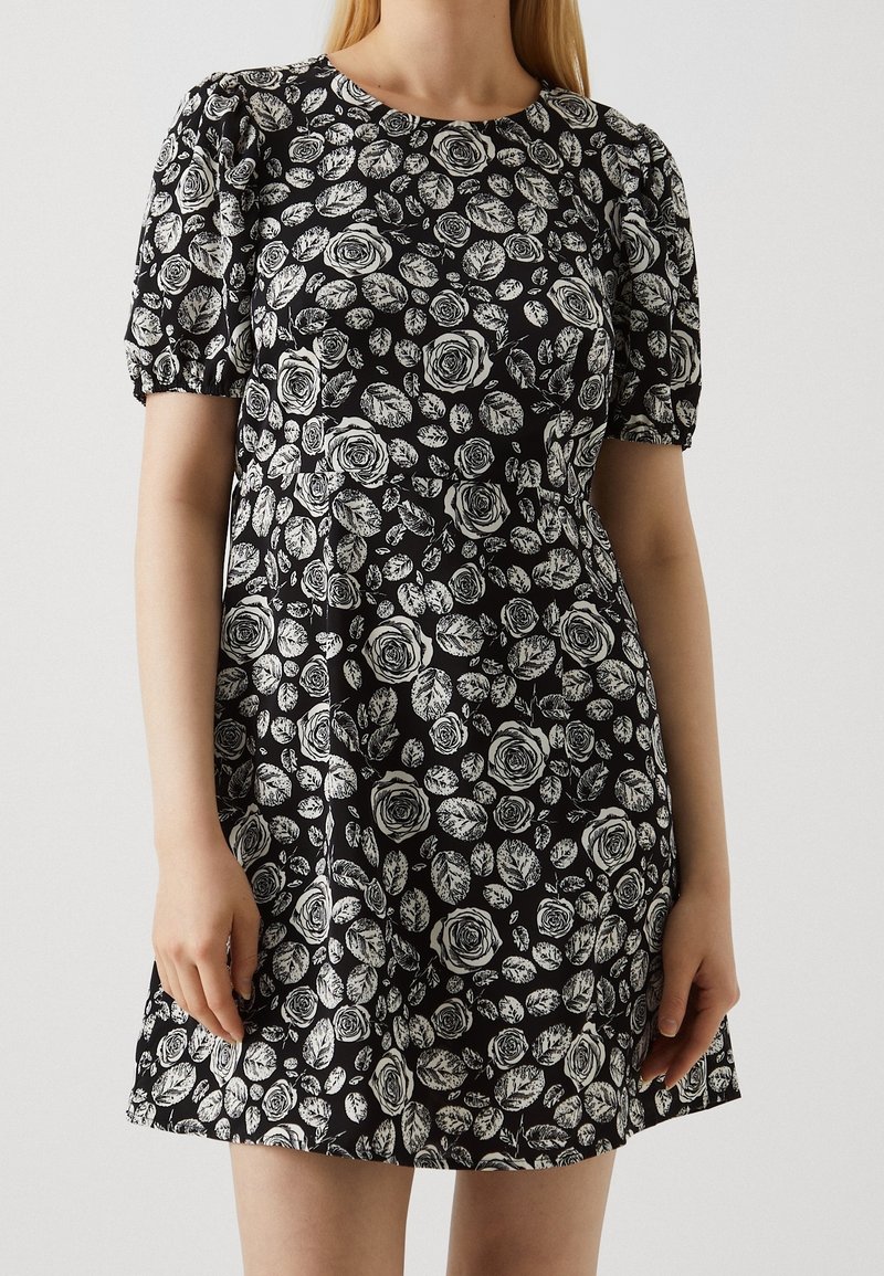 Černé šaty s bílými květinovými vzory růží a listů, krátké nafouknuté rukávy, kulatý výstřih a volný silueta.