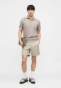 Νεαρός άνδρας με μπεζ πόλο μπλούζα, ανοιχτόχρωμα χακί σορτς, λευκές κάλτσες, μοκασίνια και κρατώντας μπεζ τσάντα χιαστί σε απλό φόντο.