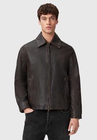 Giacca in pelle marrone con colletto appuntito e zip frontale, dotata di due tasche laterali e una finitura testurizzata. Indossata sopra jeans neri.