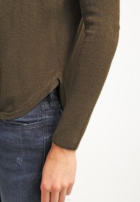 Suéter de punto verde oliva con textura acanalada, que presenta un dobladillo de corte asimétrico y mangas largas. Combinado con jeans de mezclilla oscuros, que muestran un ligero desgaste.
