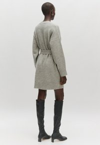 Grijze gebreide jurk met lange mouwen, ceintuur in de taille en geribbelde manchetten. Gecombineerd met zwarte leren laarzen tot de knie. Soepele textuur, casual ontwerp.