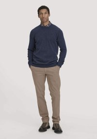 Marineblauer Strickpullover, beiges Hemd und dunkle Schuhe, getragen von einer Person, die mit den Händen in den Taschen steht, mit einem karierten Kragen.