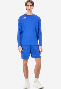 Blå träningströja med lång ärm och shorts av slät väv. Har en vit logotyp på bröstet. Bärs med vita sneakers och strumpor.