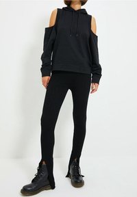 Zwarte pullover met cold shoulder design en trekkoord, gecombineerd met zwarte geribbelde leggings met zijsplitten en zwarte veterschoenen.