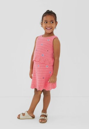 Junges Mädchen lächelt, trägt ein ärmelloses, rosa gehäkeltes Kleid mit kleinen Blumenmustern und beige Riemchensandalen, steht vor weißem Hintergrund.