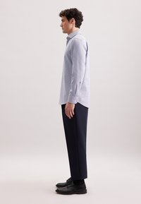 Camicia a righe blu chiaro, con bottoni e maniche lunghe, abbinata a pantaloni scuri e scarpe nere. La camicia ha un colletto classico e una forma aderente.