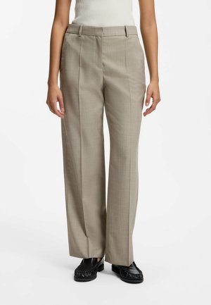 Person iført beige bukser med lige ben, hvid ærmeløs top og sorte mokkasinstil sko, stående mod hvid baggrund.
