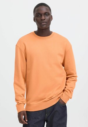 Junger Mann trägt ein einfarbiges oranges langärmeliges Sweatshirt und dunkelblaue Jeans, steht vor einem weißen Hintergrund.