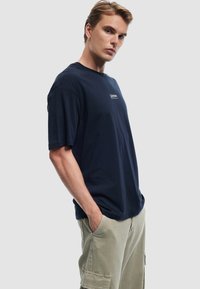 Camiseta de algodón azul marino de manga corta, con cuello redondo y un pequeño parche de logo blanco en la parte delantera. Llevada con pantalones cortos cargo en color caqui claro.