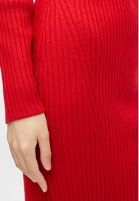 Rdeča obleka z rebrasto strukturo, ki ima teksturirano tkanino in navpične linije ter prilegajočo zasnovo; vidna je roka, ki počiva na tkanini.