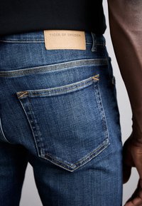 Bakifrån av en person som bär blå denimjeans med synlig bakficka och läderetikett från "Tiger of Sweden" på midjebandet.