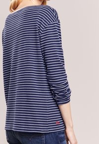 Frau trägt ein navyblaues Langarmshirt mit weißen horizontalen Streifen und rober "RL"-Stickerei am unteren Rücken, kombiniert mit blauen Jeans.