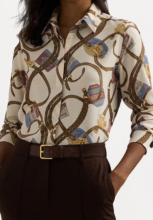Femme portant un chemisier crème avec un imprimé corde marron et étiquette de voyage, rentrée dans un pantalon taille haute marron foncé avec ceinture et mains dans les poches.