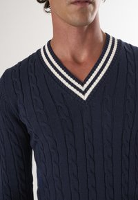 Maglione lavorato a maglia blu navy con motivo a trecce, design a V, e accenti bianchi a righe sul colletto. Tessuto testurizzato, maniche lunghe.