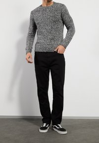 Grau gesprenkelter Strickpullover mit Rundhalsausschnitt, kombiniert mit schwarzen Hosen und schwarzen Sneakers mit weißen Akzenten und Schnürsenkeln.