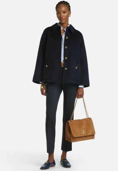 Tmavomodrý vlnený plášťový kabát s zlatými gombíkmi, dvoma prednými vreckami, v kombinácii s čiernymi nohavicami na mieru, modrými mokasínmi a hnedou semišovou kabelkou.